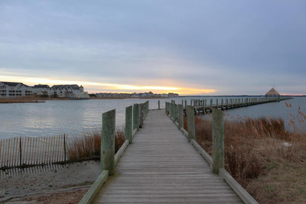 Wooden dock extending into the bay in Ocean City, Maryland, with calm water, coastal grasses, and soft sunset light.